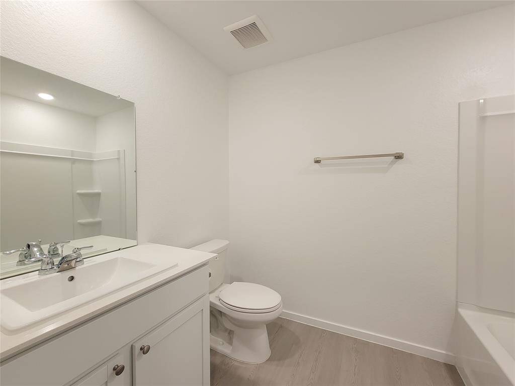 This is a clean, modern bathroom featuring a vanity with a sink and storage, a large mirror with a shelf, a toilet, and a tub with a shower space. The room has neutral colors and wood-look flooring.