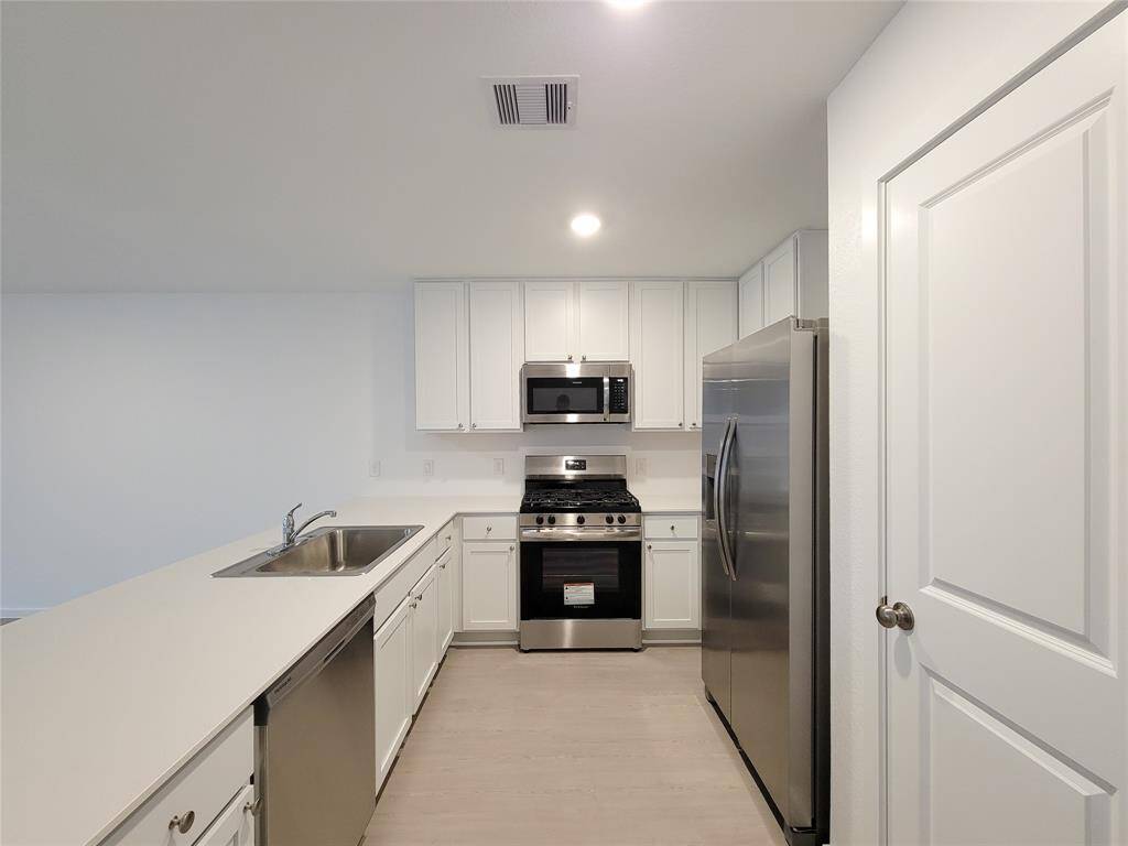 This is a modern, streamlined kitchen featuring white cabinetry, stainless steel appliances, and a neutral countertop. It has a built-in dishwasher, a range oven, and a large refrigerator, with ample storage space. The flooring is a light wood-like material, and the room is well-lit with a clean, fresh appearance.