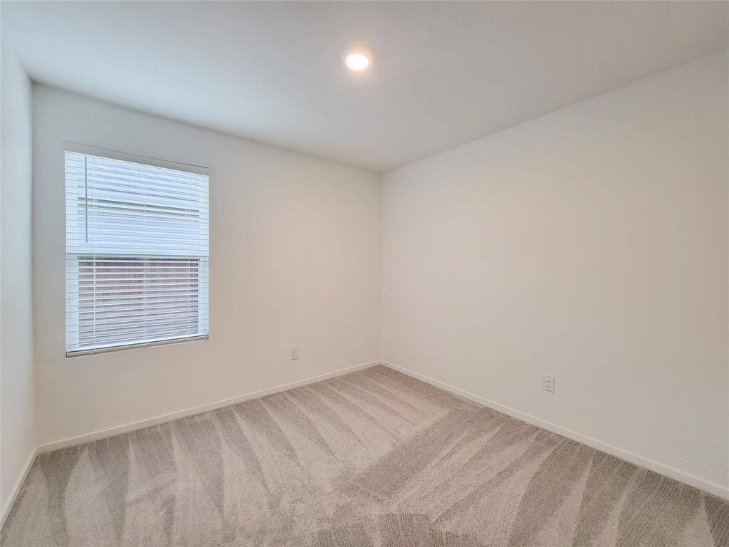 This is a bright and clean empty room featuring neutral walls, a single window with horizontal blinds, and carpeted flooring. Two ceiling lights provide ample illumination.
