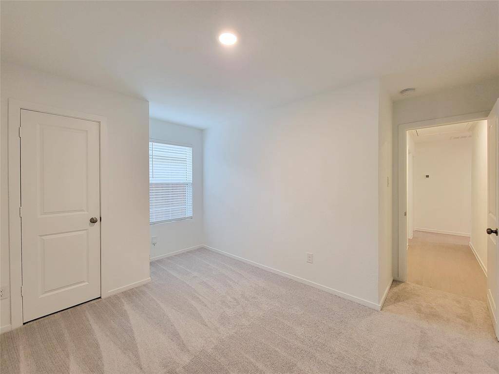 This is a bright, clean room featuring neutral-colored carpeting, fresh white walls, a window with blinds, and a closed door leading to another part of the home.