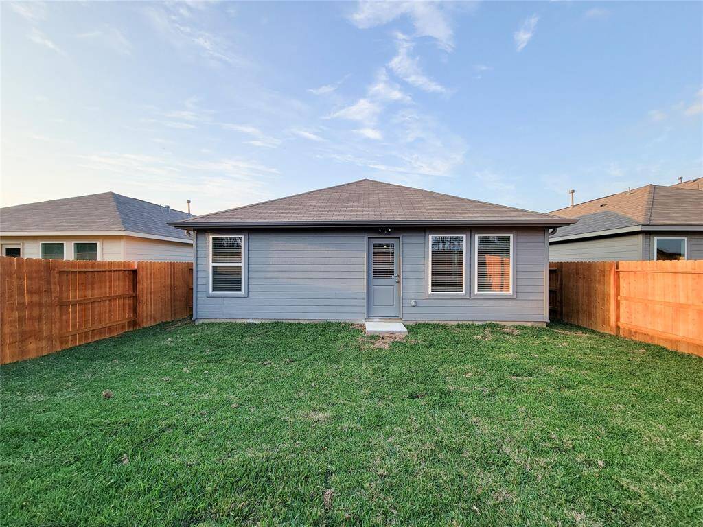 This is a photo of the rear exterior of a single-story home featuring a gray facade with white trim, two windows, and a central door. The property includes a well-maintained lawn and a wooden privacy fence.