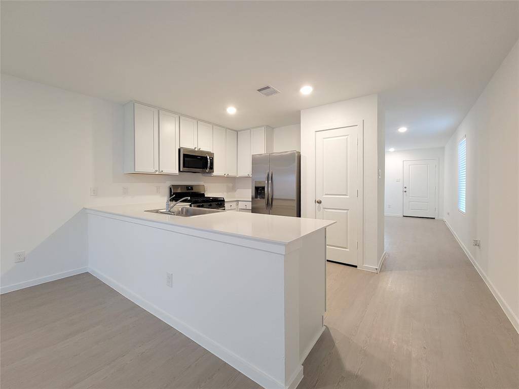 This is a modern, open-plan kitchen with white cabinetry and stainless steel appliances, featuring a central island and light wood-look flooring. The space is well-lit and leads to other rooms with a similar clean, neutral design.