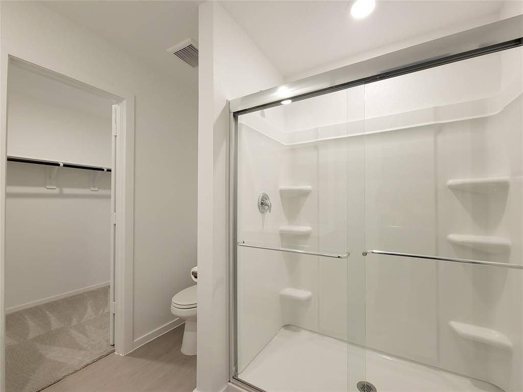 This is a modern, clean bathroom with a glass-enclosed shower, built-in shelving, a toilet, and a separate closet area with ample hanging space. The neutral color palette provides a bright and airy feel.