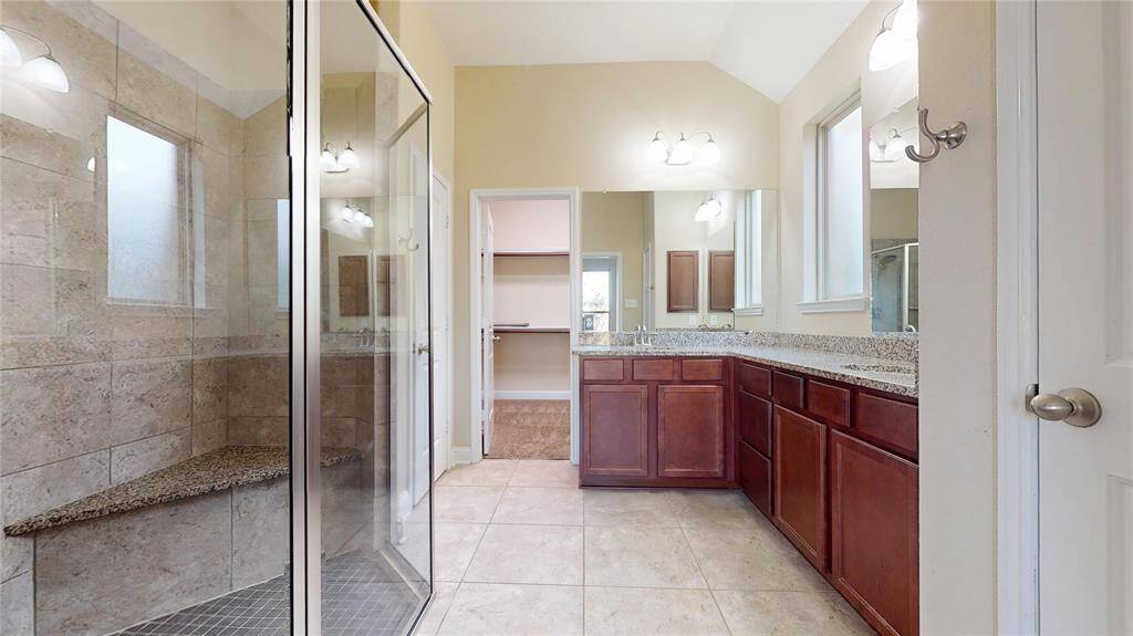 This Primary Bathroom features a spacious walk-in shower with a built-in bench, double sinks with granite countertops, and ample cabinet storage. Natural light flows through the windows, highlighting the neutral tile flooring and modern fixtures.