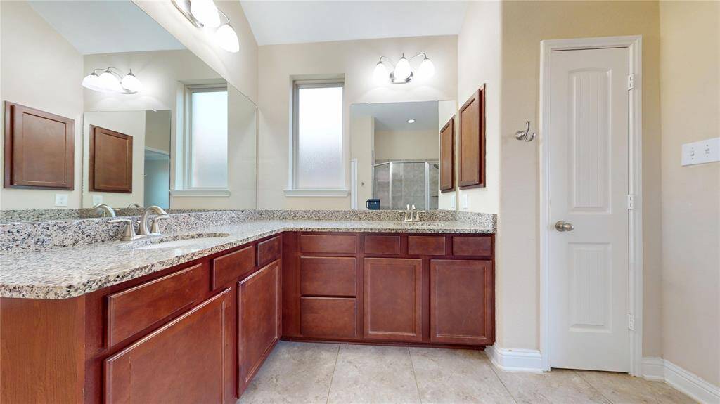 This bathroom features a spacious double vanity with granite countertops and rich wooden cabinets. It includes modern fixtures, two large mirrors, and ample natural light from two frosted windows.