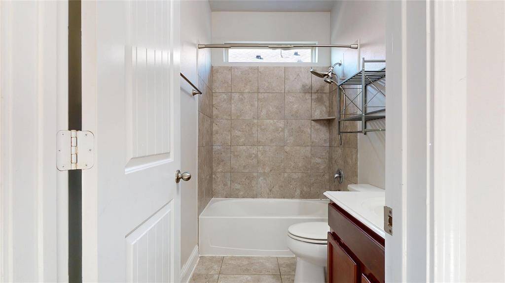 Guest bathroom featuring a tub-shower combo with beige tiles, a small window, a white vanity with a dark wood cabinet, and a wall-mounted metal shelf for storage.