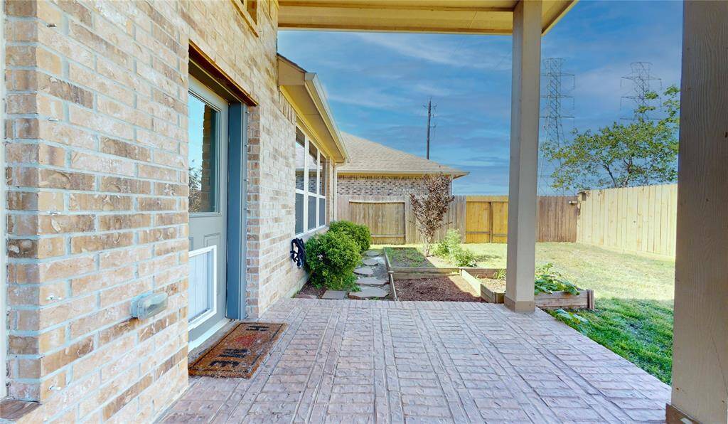This photo showcases a charming covered patio with brick flooring, leading to a fenced backyard. The space offers garden beds and a pleasant area for outdoor relaxation or entertainment.