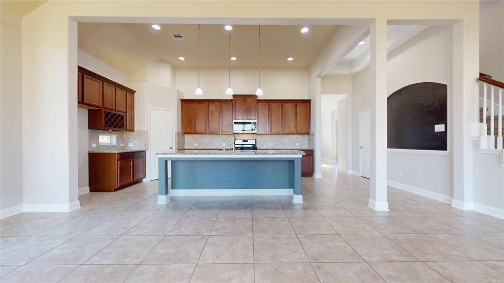 This open-concept kitchen features a spacious island with pendant lighting, granite countertops, and ample wooden cabinetry. The area is complemented by tiled floors and high ceilings, creating a bright and inviting atmosphere.