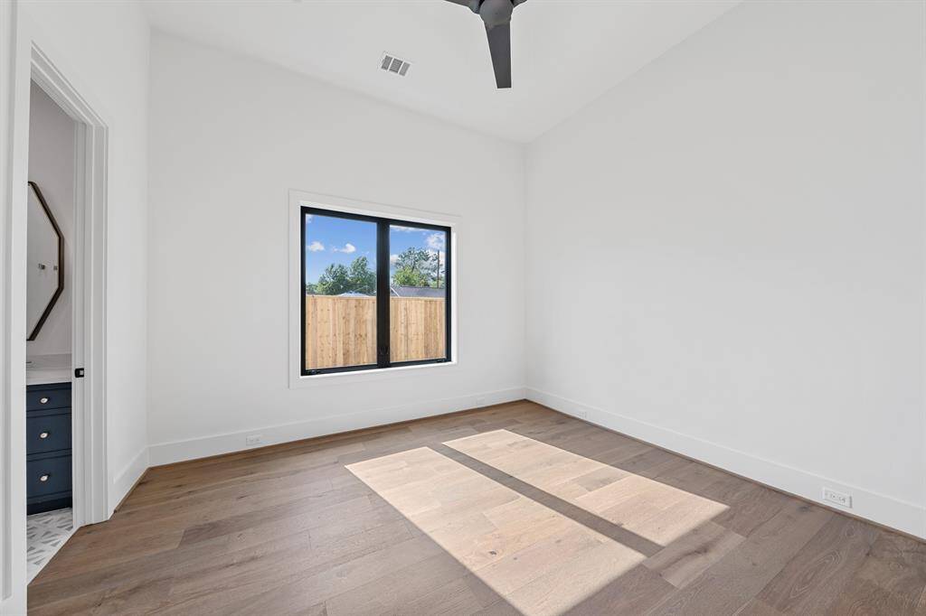 This downstairs secondary bedroom offers privacy and versatility—ideal for guests or multigenerational living. Soft natural light filters through large windows, highlighting the wide-plank oak flooring and serene neutral palette. The ensuite bath continues the refined aesthetic with designer tile, sleek fixtures, and a spacious walk-in shower.