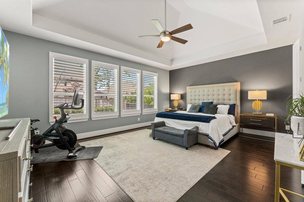 The luxurious primary suite features rich wood floors, a tray ceiling with fan, and a wall of windows with plantation shutters—creating a peaceful and private escape.