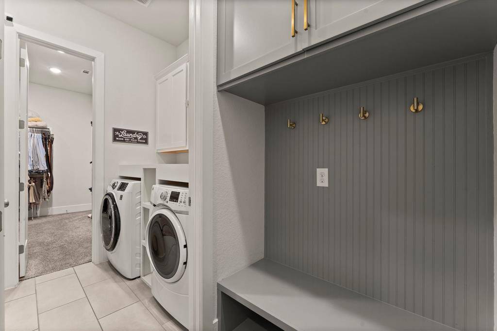 This customized laundry space includes built-in cabinetry, a folding station, and a mud area with storage hooks and bench—conveniently located off the garage and closet.