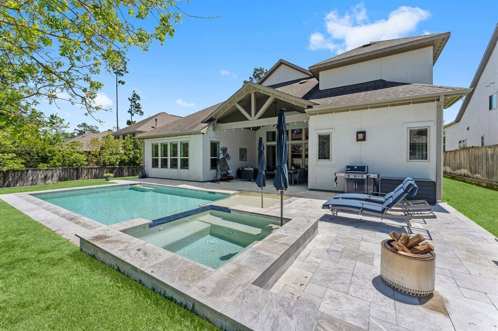 The elevated perspective captures the architectural elegance of the back of the home, showcasing the dramatic vaulted ceiling of the covered patio, the extended stone decking, and the pristine waters of the pool and spa.