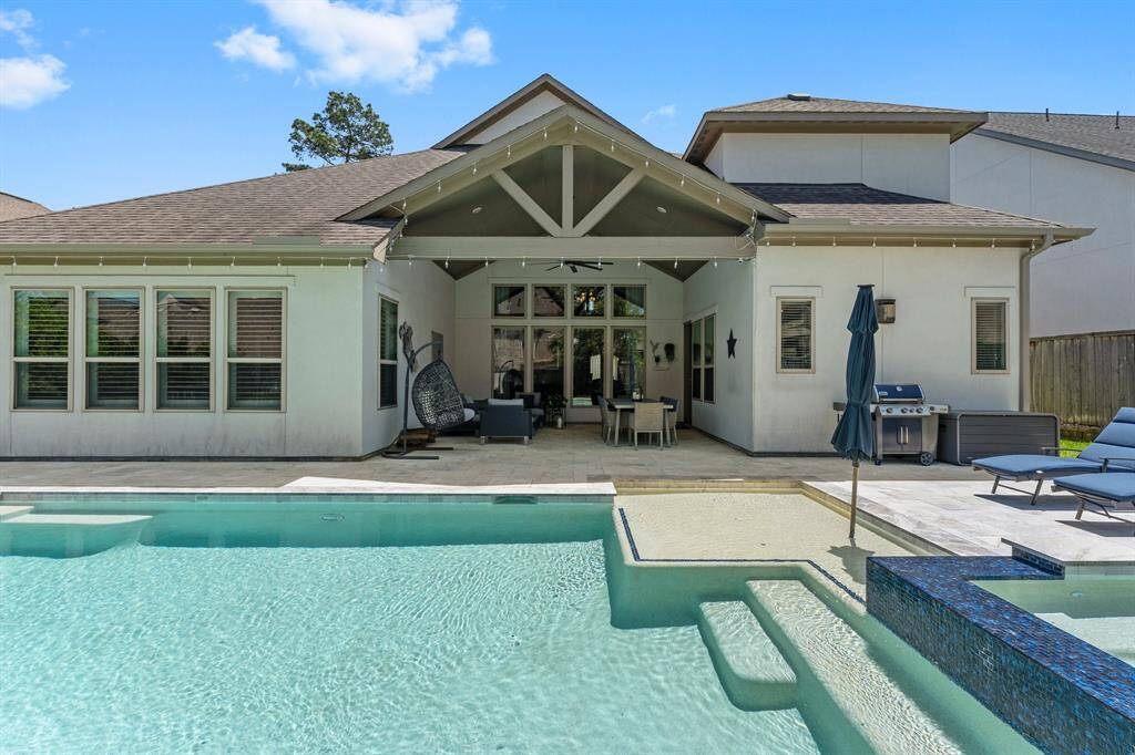 The elevated perspective captures the architectural elegance of the back of the home, showcasing the dramatic vaulted ceiling of the covered patio, the extended stone decking, and the pristine waters of the pool and spa.