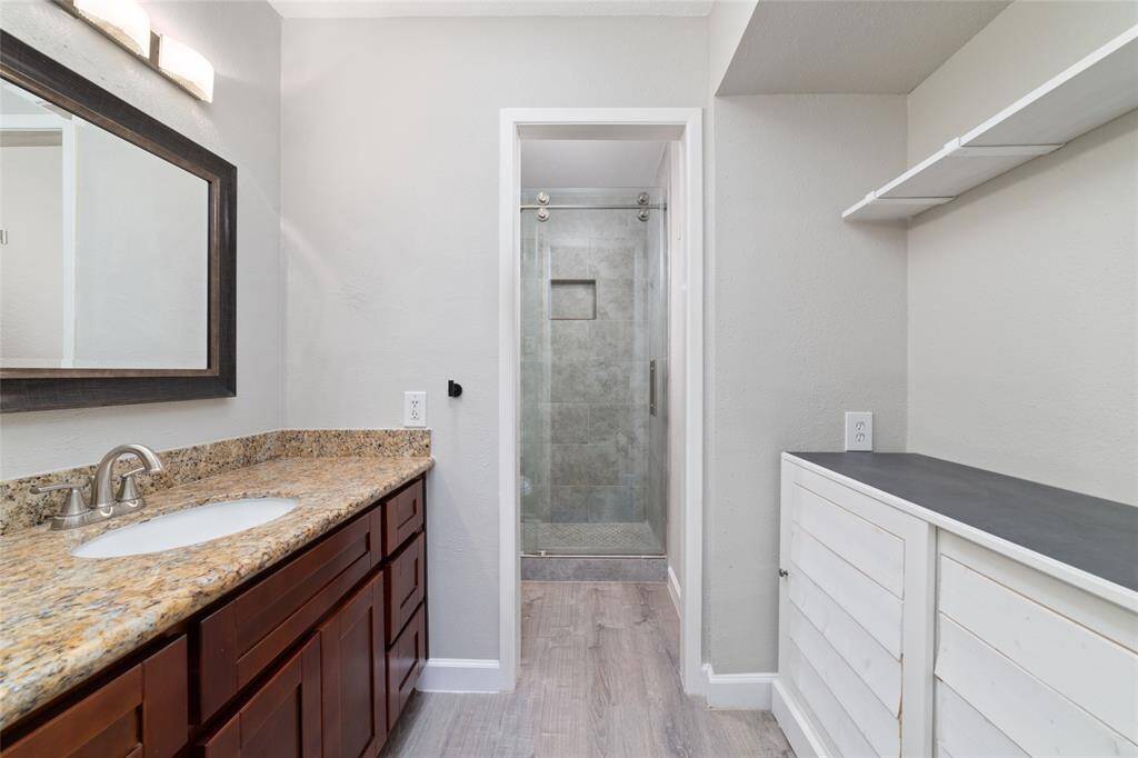 The primary bathroom features a granite countertop, dark wood cabinetry, and a large mirror, with additional storage across. A glass-enclosed shower, light gray walls, and practical storage shelves enhance the space with both style and function.