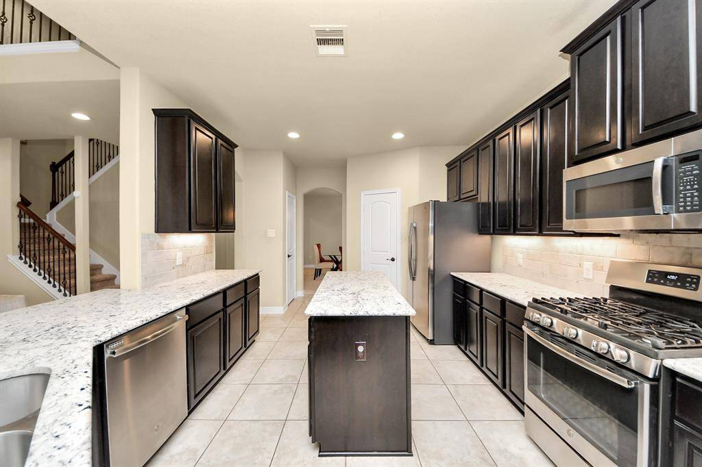 The kitchen features dark wood cabinets, light granite countertops and stainless steel appliances, including a gas stove and built-in microwave. The space is well-lit with recessed lighting, and offers a spacious layout with an island.