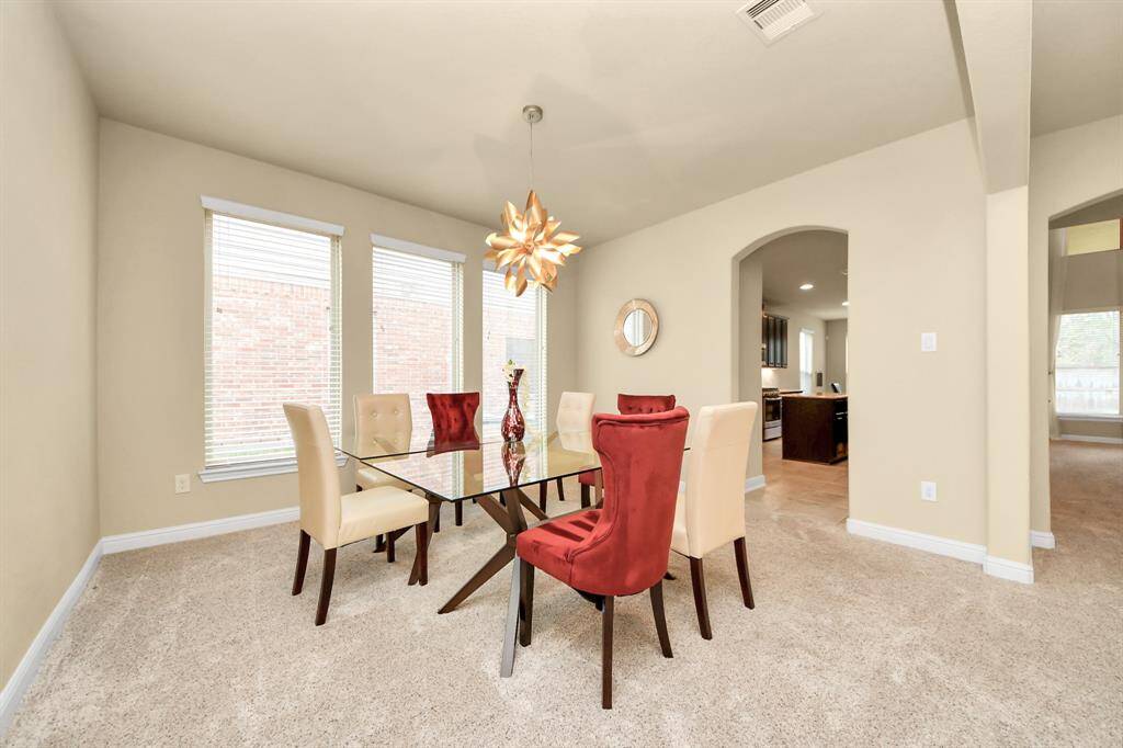 The bright dining room has a modern glass table and six chairs. The room features a unique overhead light fixture, large windows, and neutral walls create a spacious feel. The room connects to a kitchen area through an arched doorway.