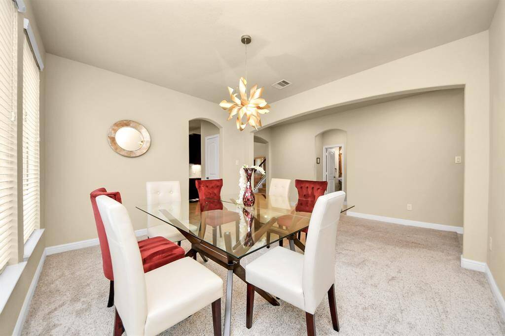 The bright dining room has a modern glass table and six chairs. The room features a unique overhead light fixture, large windows, and neutral walls create a spacious feel. The room connects to a kitchen area through an arched doorway.