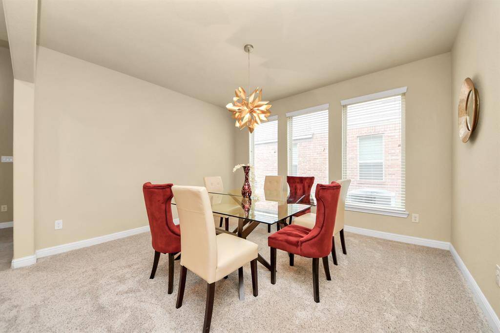 The bright dining room has a modern glass table and six chairs. The room features a unique overhead light fixture, large windows, and neutral walls create a spacious feel. The room connects to a kitchen area through an arched doorway.