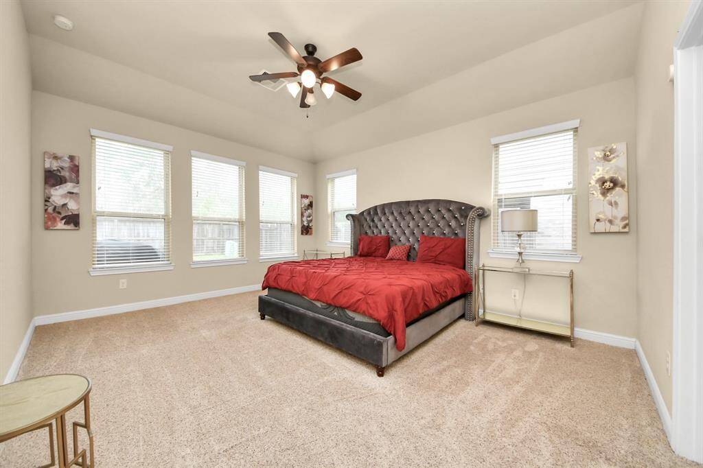The spacious, first-floor primary bedroom has plush carpet and a luxurious king-size bed with a tufted headboard. The room is bright with multiple windows and has a ceiling fan for comfort.