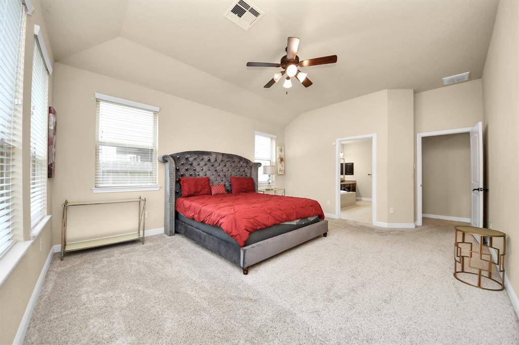 The spacious, first-floor primary bedroom has plush carpet and a luxurious king-size bed with a tufted headboard. The room is bright with multiple windows and has a ceiling fan for comfort.