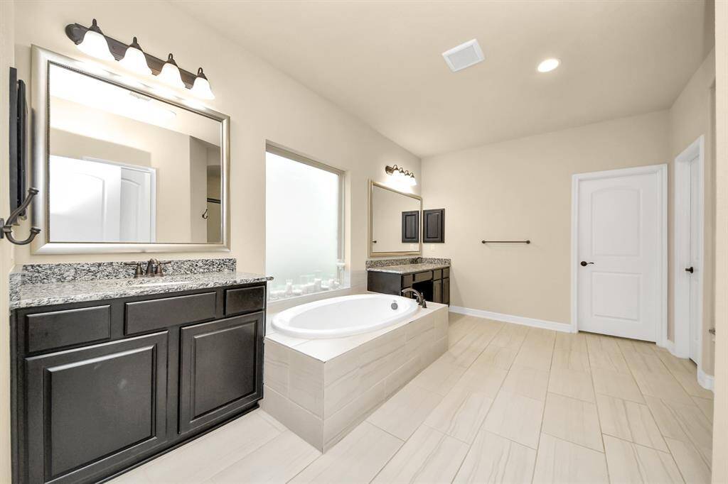 This primary bathroom features a modern design with dual vanities, granite countertops, a large soaking tub, and ample natural light. The neutral tones and sleek fixtures create a spa-like atmosphere.