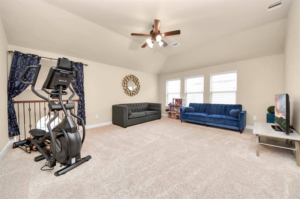 The game room invites ample natural light from three large windows and there is a ceiling fan for comfort. It includes two modern sofas, a TV, and exercise equipment, offering a versatile space for relaxation and fitness.