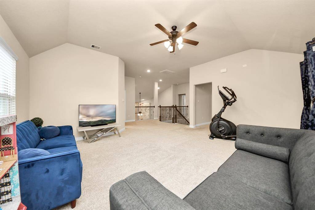 The game room invites ample natural light from three large windows and there is a ceiling fan for comfort. It includes two modern sofas, a TV, and exercise equipment, offering a versatile space for relaxation and fitness.