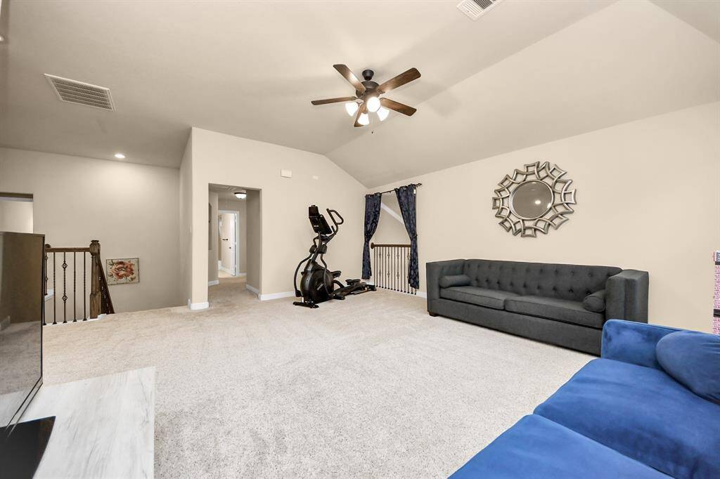 The game room invites ample natural light from three large windows and there is a ceiling fan for comfort. It includes two modern sofas, a TV, and exercise equipment, offering a versatile space for relaxation and fitness.