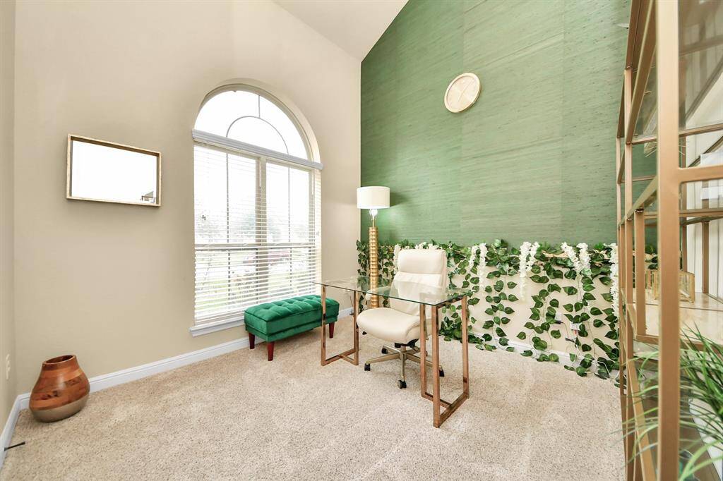 The Office features a high ceiling and large arched window for natural light. It has a modern, glass-top desk, a green accent wall with decorative greenery, and a plush carpet, creating a cozy yet stylish workspace.