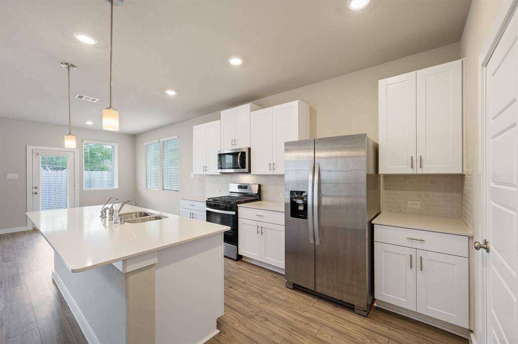 This kitchen features modern stainless steel appliances, white cabinetry, and a spacious island with a double sink. The open layout is complemented by recessed lighting and natural light from large windows and a glass door.