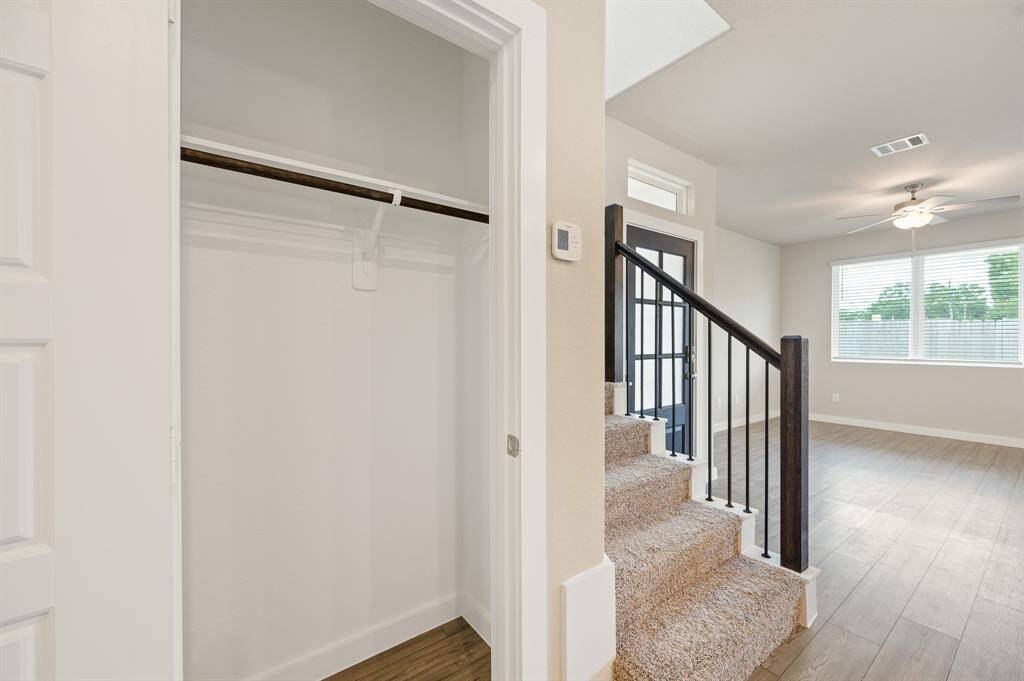 This photo shows a bright, modern entryway with a small carpeted staircase leading to an upper level. Adjacent is a spacious open closet with a hanging rod. The room has large windows, wood-style flooring, and a ceiling fan, creating an inviting atmosphere.