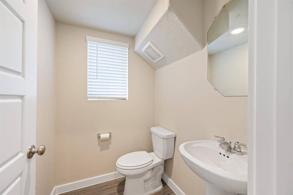 This is a compact, well-lit half bathroom featuring a toilet and pedestal sink. It has a small window with blinds and a wall-mounted mirror. The neutral tones and clean lines create a modern, functional space.