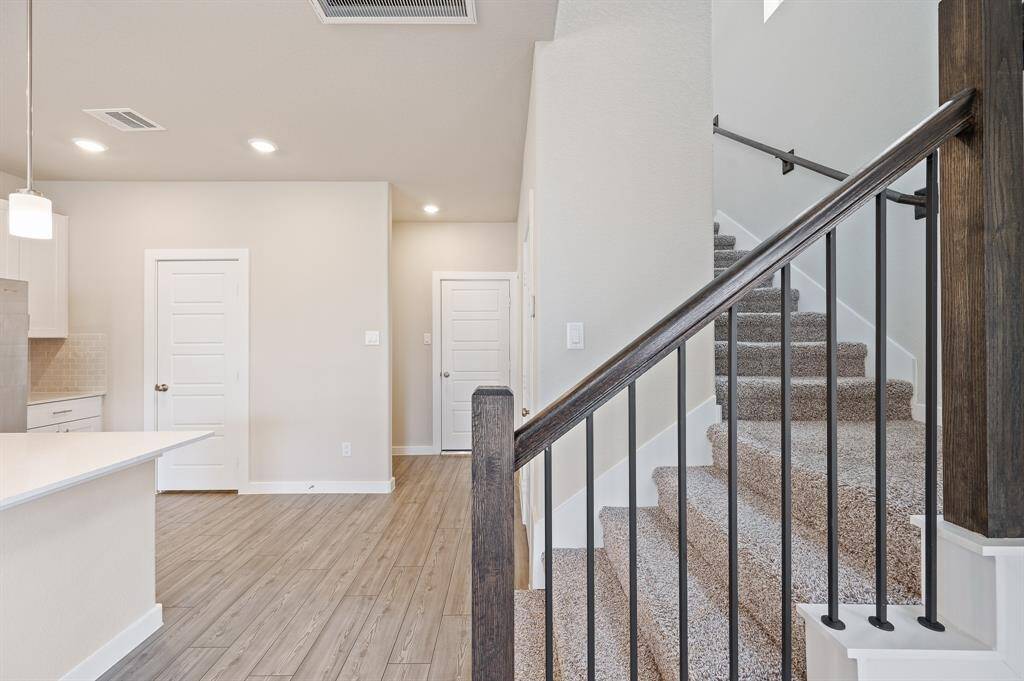 This photo showcases a modern interior with a carpeted staircase featuring a sleek black railing. The area has light wood style flooring and neutral walls, complemented by recessed lighting and a stylish pendant light over the kitchen counter. The overall look is clean and contemporary.