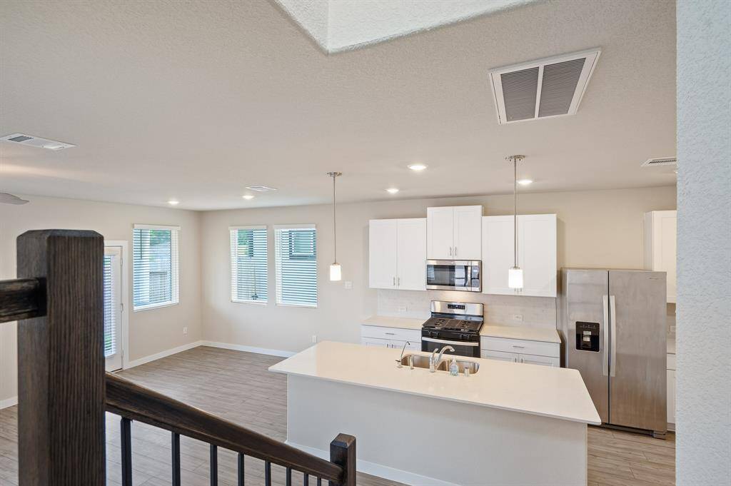 This modern kitchen features sleek white cabinetry, stainless steel appliances, and a spacious island with a sink. It opens to a bright living area with ample natural light from large windows and recessed lighting for a contemporary feel.