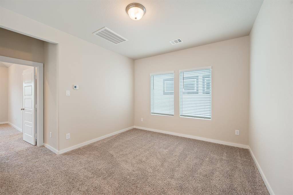 A bright, carpeted room with neutral walls, featuring two windows with blinds and a ceiling light. There's an open doorway leading to another space, enhancing the room's spacious feel.