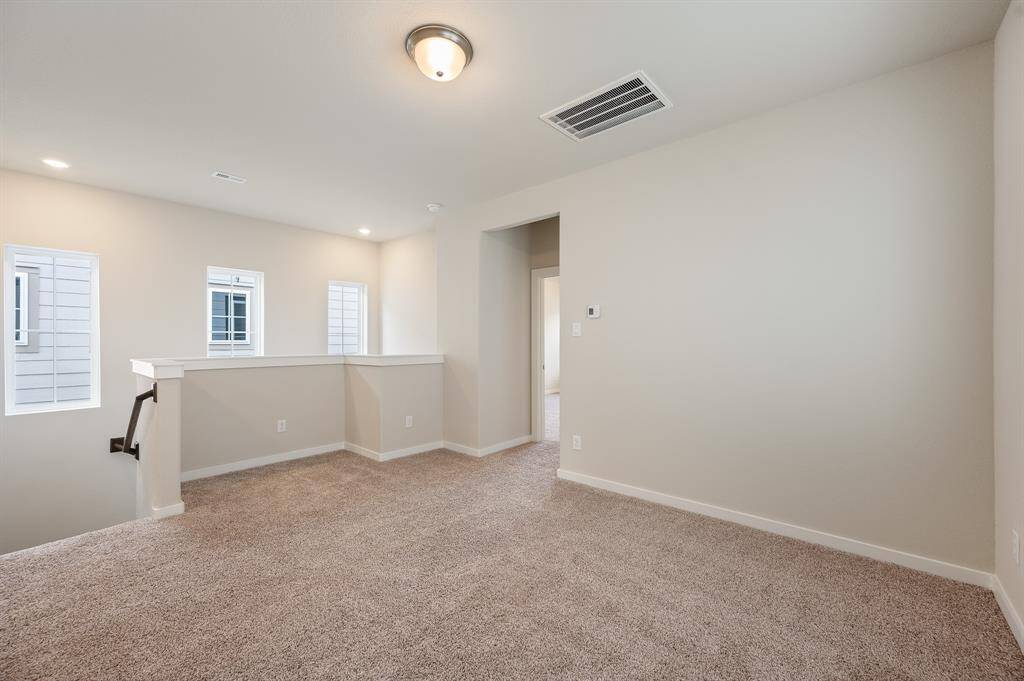 This is a bright, carpeted room with neutral walls and a recessed ceiling light. It features three windows for natural light, a short railing overlooking a staircase, and an open doorway leading to another room.