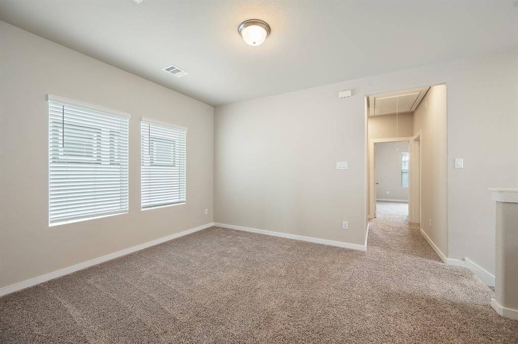 This photo shows a bright, carpeted room with neutral walls, featuring two windows with blinds. It has a ceiling light fixture and an open doorway leading to another room, offering a cozy and inviting space.