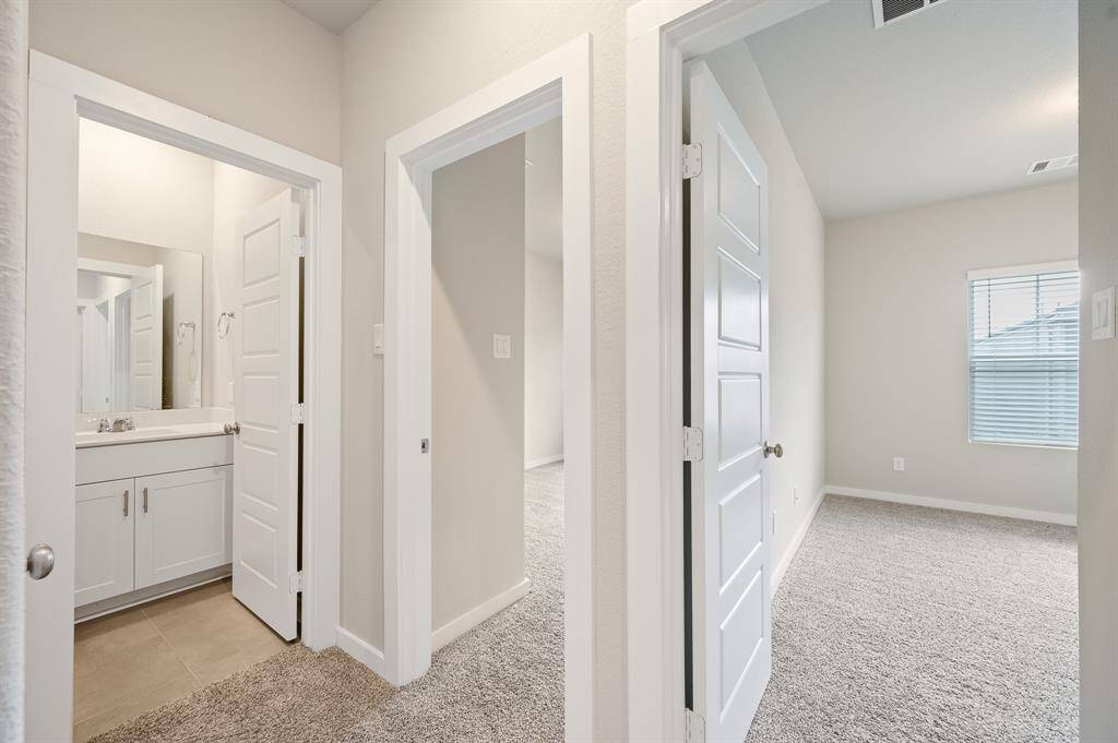 The photo shows a bright, modern hallway with carpeted flooring leading to a bathroom and a bedroom. The bathroom has a white vanity and tiled floor, while the bedroom features a large window for natural light. Fresh, neutral tones enhance the spacious feel.