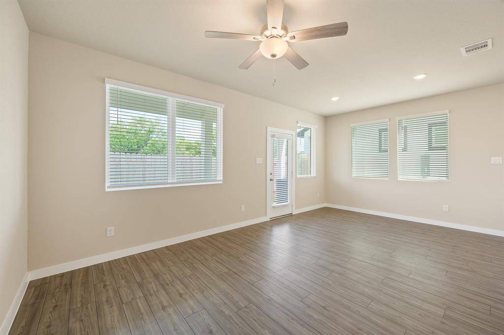 This spacious, light-filled room features wood-look flooring, neutral walls, and a ceiling fan. Large windows with blinds provide ample natural light, and a glass door leads to the outside, enhancing indoor-outdoor flow.