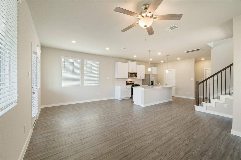 This spacious open-concept living area features modern wood style floors, recessed lighting, and a ceiling fan. The kitchen boasts stainless steel appliances, white cabinetry, and an island with pendant lights. A staircase with sleek railing leads to the upper level, and large windows provide ample natural light.