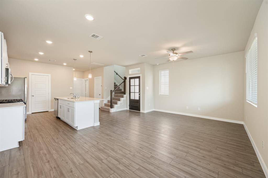 The photo showcases a spacious, open-concept living area with light wood style flooring, neutral walls, and recessed lighting. The modern kitchen features a large island, stainless steel appliances, and pendant lights. A staircase leads to the upper level, and there's ample natural light from several windows.