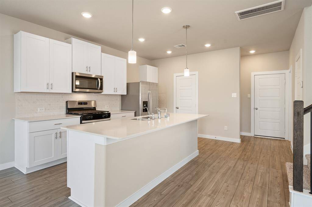 This modern kitchen features sleek white cabinetry, stainless steel appliances, and a spacious island with a sink. The open layout is enhanced by wood-style flooring and recessed lighting, providing a bright and welcoming atmosphere.