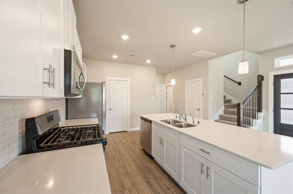 Modern kitchen with white cabinetry, stainless steel appliances, and a large island with a sink. It features recessed lighting, wood style flooring, and an open layout leading to a staircase.