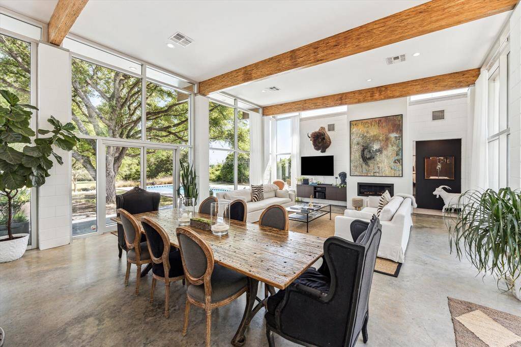 Step into this spacious, modern living and dining area, where large windows flood the room with natural light, creating a warm and inviting atmosphere. The exposed wooden beams on the ceiling add a touch of rustic charm, perfectly complementing the contemporary polished concrete floor.