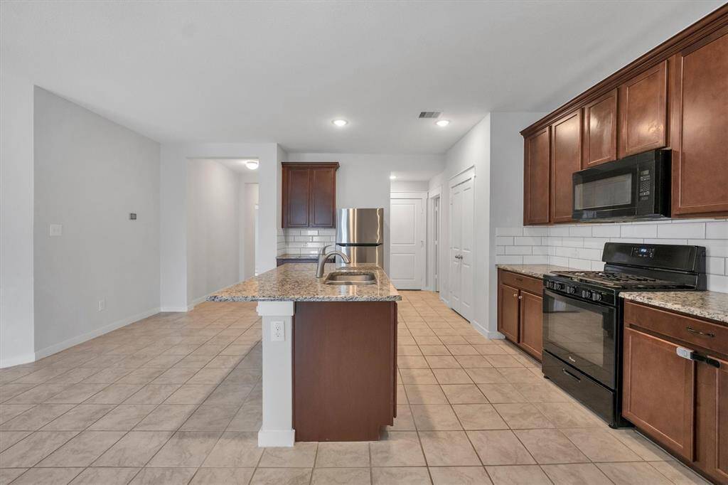 This kitchen features a spacious pantry with white double doors, granite countertops, dark wood cabinets, and modern appliances including a black stove and microwave. The tile floor complements the sleek, tiled backsplash.