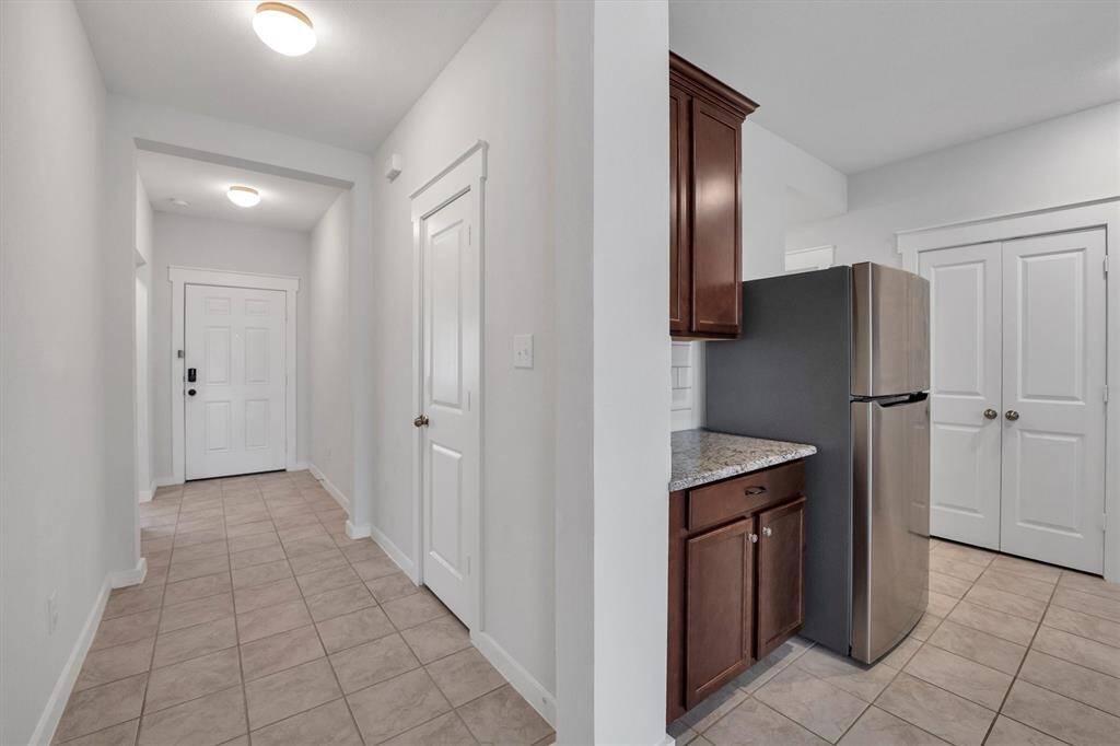 This photo shows a bright, tiled hallway leading to a kitchen area. The kitchen features stainless steel refrigerator that is included, granite countertops, and dark wood cabinets, providing a clean and modern look.
