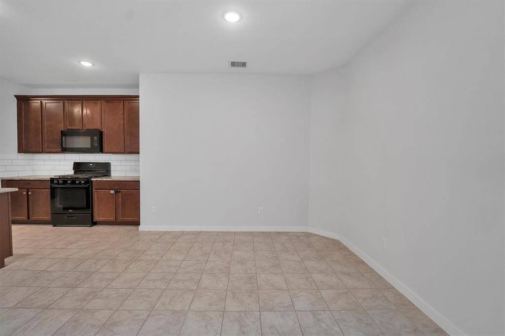 This photo shows a modern kitchen area with brown cabinets, a black stove, and microwave. It features a tiled floor and white subway tile backsplash, offering a clean, spacious look.