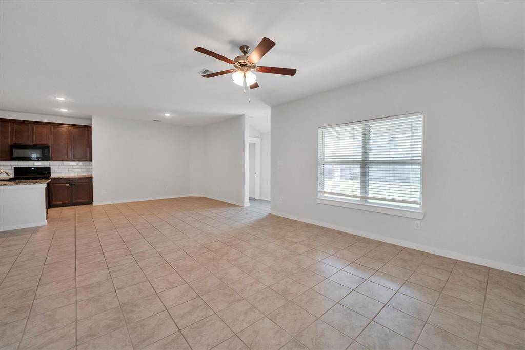 This spacious living area features neutral tile flooring, ample natural light from multiple windows, and a ceiling fan for comfort. An open doorway leads to an adjoining room, offering versatile space for various uses.