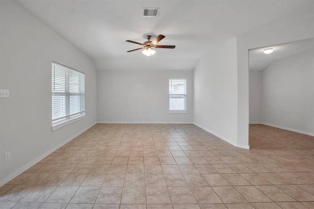 This spacious living area features tile flooring, a ceiling fan, and ample natural light from a large window. It seamlessly connects to a modern kitchen with dark cabinetry and a tiled backsplash.