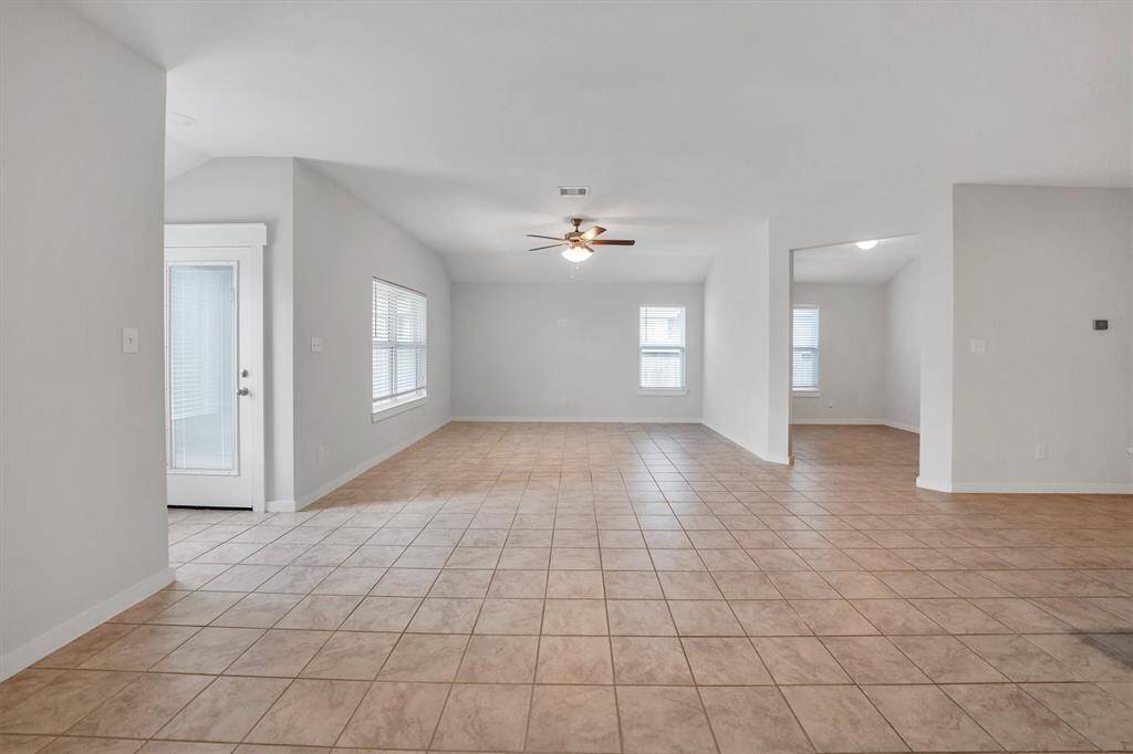Spacious open-concept living area with neutral tile flooring, ample natural light from multiple windows, and a ceiling fan. Adjacent flex room accessible through wide doorway, and a door leading to the outside enhances accessibility.